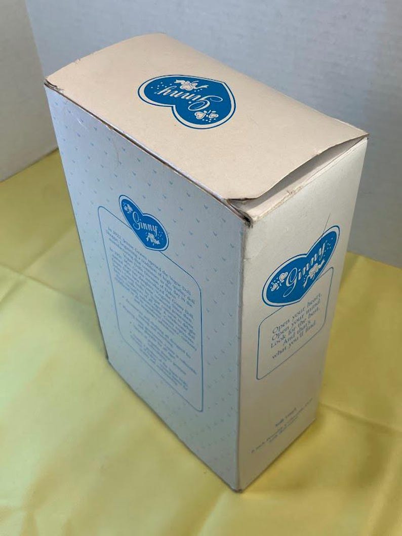 Puede incluir: Una caja de color crema con logotipos azules en forma de coraz&oacute;n floral y el nombre "Ginny". La caja tiene el texto "Abre tu coraz&oacute;n, Abre tu mente, Busca lo mejor, Y lo que encontrar&aacute;s."