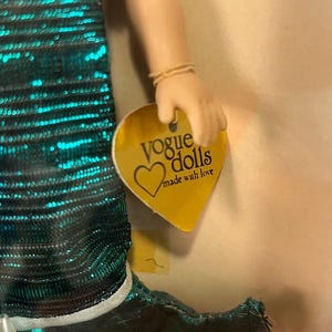 Puede incluir: Una mu&ntilde;eca con un vestido de lentejuelas color teal con ribete blanco. La mano de la mu&ntilde;eca sostiene una etiqueta dorada que dice "Vogue Dolls made with love". La piel de la mu&ntilde;eca es clara.