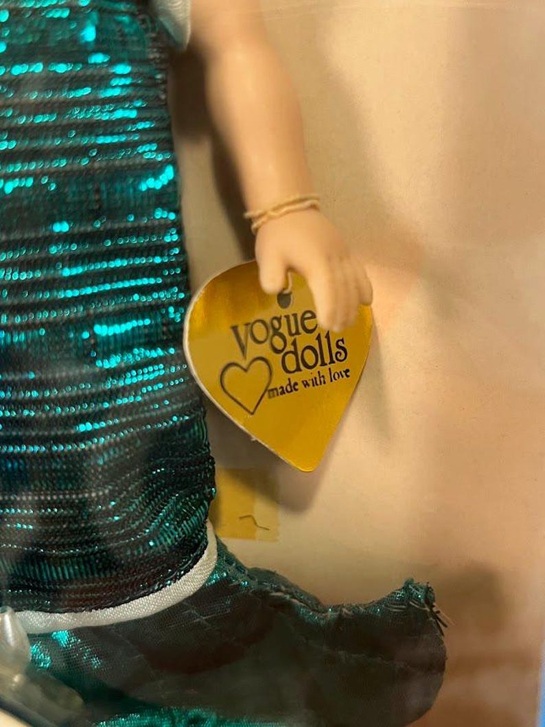 Puede incluir: Una mu&ntilde;eca con un vestido de lentejuelas color teal con ribete blanco. La mano de la mu&ntilde;eca sostiene una etiqueta dorada que dice "Vogue Dolls made with love". La piel de la mu&ntilde;eca es clara.