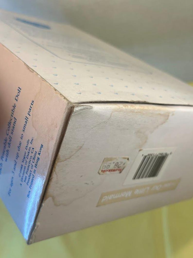 Puede incluir: Una caja de mu&ntilde;eca coleccionable vintage, con el texto "Our Little Mermaid". La caja es de color blanquecino con un patr&oacute;n de puntos azul claro. La caja tiene una etiqueta de precio y un c&oacute;digo de barras. La caja est&aacute; ligeramente da&ntilde;ada.