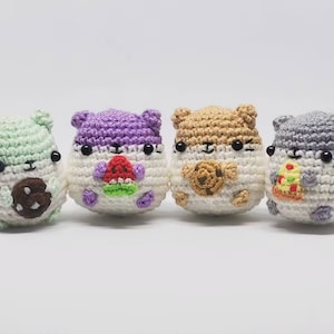 Puede incluir: Cuatro peluches de hámster de crochet, cada uno sosteniendo un alimento diferente. Los hámsters son verde, morado, marrón y gris.