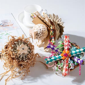 Può includere: Una collezione di giocattoli per uccelli in varie forme e consistenze. I giocattoli includono una palla intrecciata, un giocattolo a forma di girasole con carta sminuzzata e un giocattolo intrecciato colorato con un accento floreale. I giocattoli sono progettati per gli uccelli.