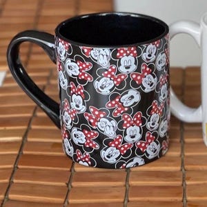 May include: Black ceramic mug featuring a pattern of Minnie Mouse faces and red polka dot bows. The mug has a black handle and interior. A white mug is partially visible in the background.