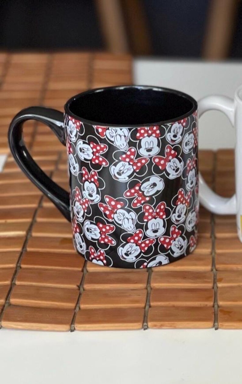 May include: Black ceramic mug featuring a pattern of Minnie Mouse faces and red polka dot bows. The mug has a black handle and interior. A white mug is partially visible in the background.