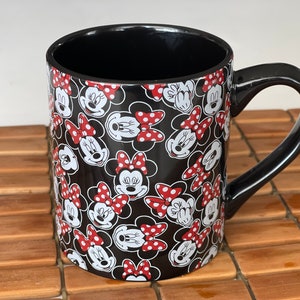 May include: Black ceramic mug featuring a repeating pattern of Minnie Mouse faces with red polka dot bows on a black background. The mug has a black interior and a curved black handle. The mug is sitting on a wooden surface.