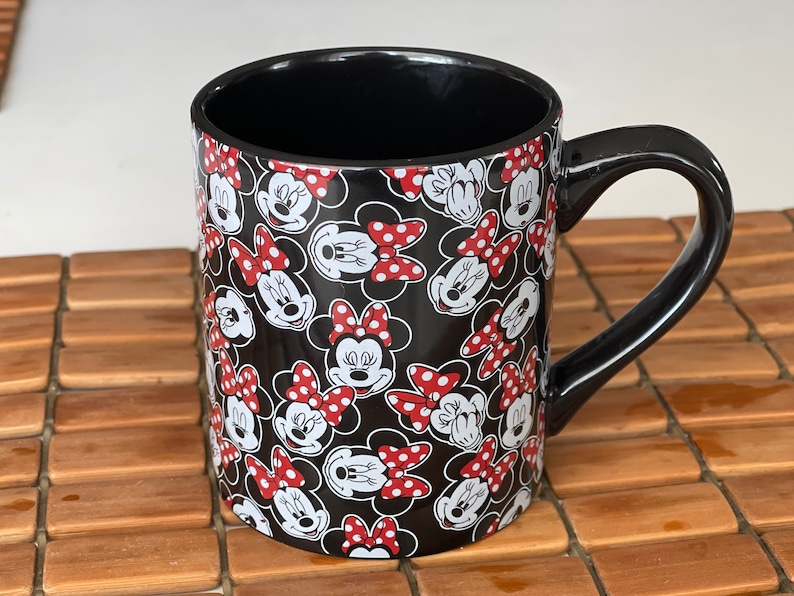 May include: Black ceramic mug featuring a repeating pattern of Minnie Mouse faces with red polka dot bows on a black background. The mug has a black interior and a curved black handle. The mug is sitting on a wooden surface.
