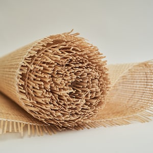 May include: A roll of natural brown woven cane webbing with fringed edges. The webbing is rolled up tightly and the fringe is visible on both ends.