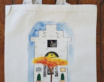 Puede incluir: Bolso tote color crema con una ilustración en acuarela de un edificio con un árbol y el texto "shepherdstown, wv". El edificio tiene un diseño de sol sobre el árbol y el texto "S 2". El bolso tiene asas beige.
