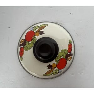May include: A round, off-white pot lid with a black handle. The lid is decorated with colorful illustrations of vegetables, including tomatoes, carrots, and corn. The lid has a silver rim.