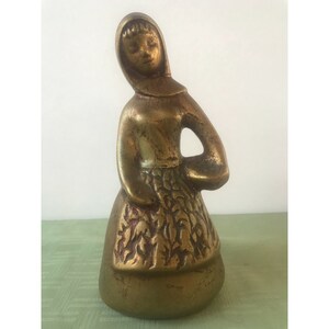 May include: A gold-toned metal figurine of a woman wearing a long dress and a hooded cloak. The dress has a detailed floral pattern.