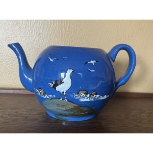 May include: A blue ceramic teapot with a white seagull and waves painted on the side. The teapot has a spout and a handle.