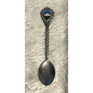 May include: Silver-toned souvenir spoon with a twisted handle and a decorative top featuring a shield design with the words "Luray Caverns" and a depiction of a building. The spoon's bowl has an etched image of a building.