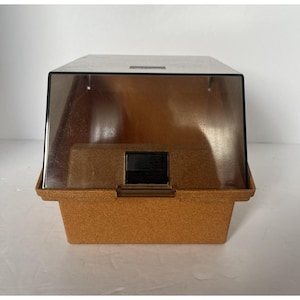 May include: A vintage brown plastic cassette tape storage case with a clear, hinged lid. The case has a rectangular shape with a textured surface and a black latch. The top is a darker brown color.