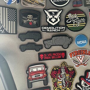 May include: A selection of embroidered patches and decals. Notable designs include a red Toyota Land Cruiser, a skull with crossed guns, and a patch with the text "Demolition Ranch". Other patches feature the text "Gryffindor" and "5.11 Tactical Series".