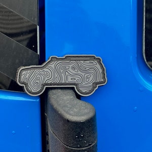May include: A grey and black topographical map design car emblem. The emblem is in the shape of a vehicle and is attached to a blue car door. The emblem has a detailed, layered design.