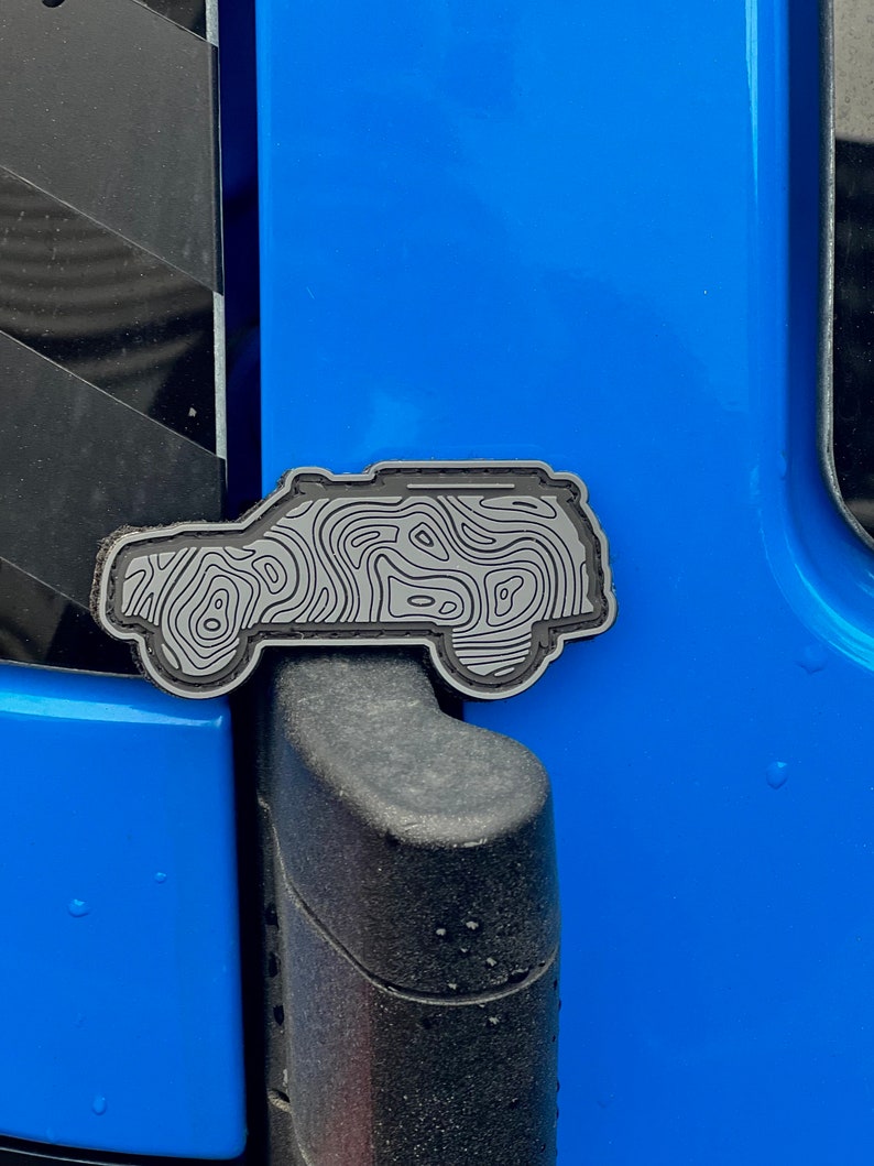 May include: A grey and black topographical map design car emblem. The emblem is in the shape of a vehicle and is attached to a blue car door. The emblem has a detailed, layered design.