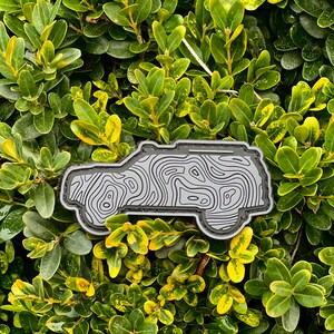 May include: A gray and black patch in the shape of a vehicle, featuring a topographic map design. The patch is set against a backdrop of green and yellow foliage.