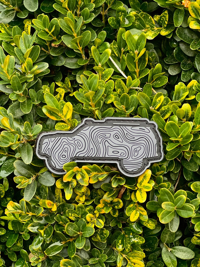 May include: A grey and black patch in the shape of a vehicle, with a topographic map design. The patch is set against a backdrop of green and yellow foliage.