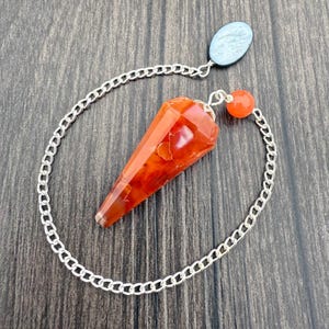 May include: A faceted, orange-red carnelian pendulum with a silver chain and a small orange bead. The pendulum is suspended from a light blue oval-shaped stone. The chain is attached to the pendulum and the oval stone.