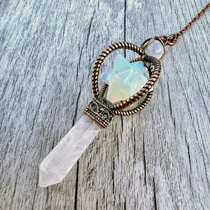 May include: A crystal pendulum with a clear quartz point and a decorative copper-colored metal setting. The pendulum features a star-shaped opalite stone and a twisted rope design. The chain is also copper-colored, and the background is a wooden surface.