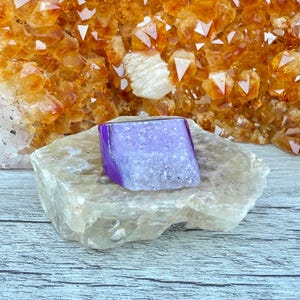 May include: A vibrant purple and white crystal geode sits atop a light-colored stone, set against a backdrop of golden-orange crystals. The geode has a smooth, polished surface with a crystalline interior, showcasing natural beauty.