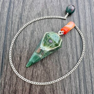 Puede incluir: Un péndulo de cristal verde, marrón y blanco con una cadena plateada y una cuenta de cornalina roja. El péndulo tiene una forma puntiaguda y facetada.