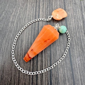 May include: An orange crystal pendulum with a silver chain and two accent stones. The main crystal is a faceted cone shape. One accent stone is a small, round, light green bead, and the other is a small, irregular orange stone.