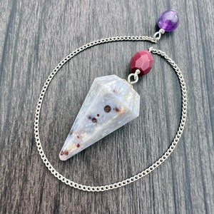 Puede incluir: Un péndulo de cadena plateada con una punta de cristal blanca y gris, una cuenta de piedra roja y una cuenta de amatista púrpura.