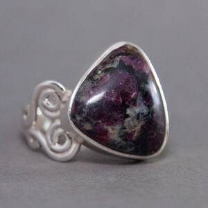 Puede incluir: Un anillo de plata con una piedra triangular de color púrpura oscuro y negro. El anillo tiene un diseño delicado y en espiral.
