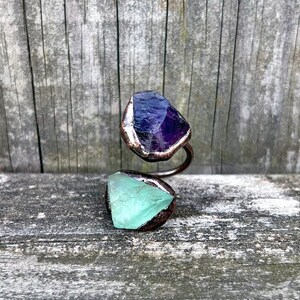 May include: A copper ring with a purple amethyst and a green fluorite stone. The ring is wrapped around a wooden surface.