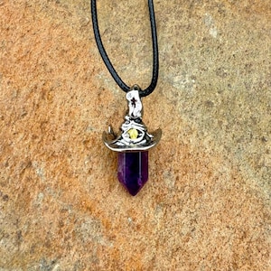 May include: A pendant necklace featuring a silver witch hat charm with a yellow gem and a deep purple crystal point. The necklace hangs on a black cord, perfect for adding a touch of mystical style to any outfit.