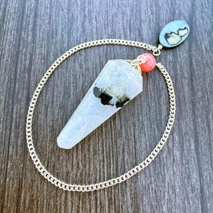May include: A pendulum with a clear, faceted crystal point, accented with black inclusions. It hangs from a silver chain, connected to a small, oval-shaped stone and a coral-colored bead. The design is set against a dark wood grain background.