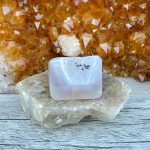 May include: A light pink and white crystal geode sits on a light grey stone, with a background of orange crystals. The geode is square-shaped with a white interior and small black inclusions.