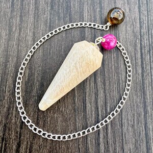May include: A beige crystal pendulum with a silver chain, a brown sphere, and a pink sphere. The pendulum is cone-shaped and has a textured surface. The chain is made of small, linked metal pieces.