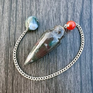 May include: A pendulum with a dark, speckled stone body and a silver chain. The pendulum has a rounded stone at the top and a red bead near the chain's end. The stone body is dark with green and brown hues.