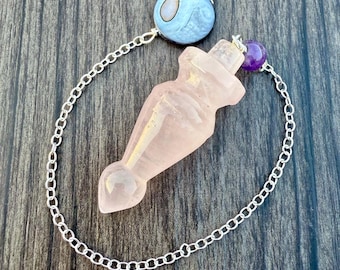 May include: A pink quartz crystal pendant in the shape of a potion bottle, connected to a silver chain. The chain is adorned with a blue and white bead and a purple bead. The pendant is on a wooden surface.