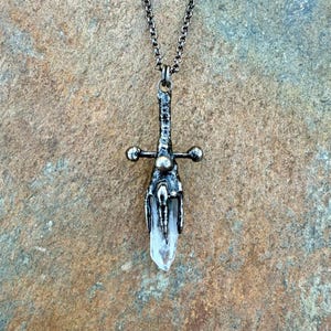 May include: A silver-toned pendant necklace featuring a sword design with a clear crystal blade. The sword has a detailed hilt and crossguard, and hangs from a delicate chain. The necklace is set against a textured, neutral-toned background.