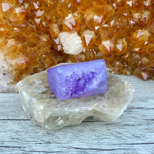 May include: A rectangular, lavender crystal geode rests on a light grey stone, with a backdrop of golden-orange crystals. The geode features a hollow centre filled with darker purple crystals. The background crystals have sharp, pointed edges.