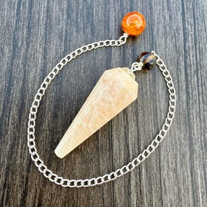 May include: A crystal pendulum with a light orange, faceted crystal point. The pendulum is attached to a silver chain, with an orange and a dark brown bead at the top. The pendulum is designed for divination and spiritual practices.