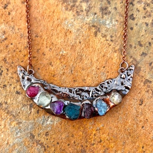 May include: A handcrafted necklace featuring a silver-toned, curved pendant with a textured design. The pendant is adorned with a row of colorful gemstones, including red, green, purple, and blue stones. The necklace has a copper-colored chain.