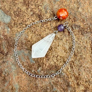 May include: A clear quartz crystal pendulum with a silver chain, an orange and red bead, and a purple bead. The pendulum is diamond-shaped and hangs from the chain. The chain is looped around the pendulum.