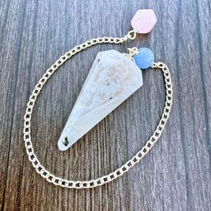 Könnte beinhalten: Ein klares Quarz-Kristall-Pendel mit einer silbernen Kette, einem rosa sechseckigen Kristall und einer blauen Kugel. Das Pendel liegt auf einer dunklen Holzoberfläche.