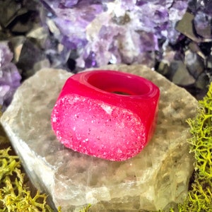 May include: A pink ring made of resin with a sparkly, glittery finish. The ring is shaped like a rounded rectangle with a smooth, polished surface.