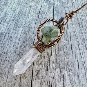 Op de afbeelding: Een heldere kwartskristallen hanger met een koperkleurige metalen setting en ketting. De hanger heeft een gesneden groene steen en een decoratieve metalen ring. Het kristal is ongeveer 7.6 cm lang.
