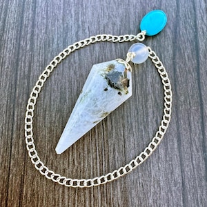 May include: A crystal pendulum with a silver chain, a blue-green stone, and a white and gray crystal point. The pendulum is designed for divination and spiritual practices. The chain is approximately 8 inches long.