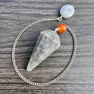 Puede incluir: Un péndulo de cristal gris con una cadena plateada y una cuenta blanca y naranja. El péndulo está sobre una superficie de madera.