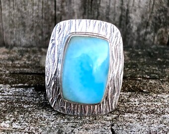 Può includere: Un anello d'argento con una pietra preziosa rettangolare azzurra. L'anello presenta un motivo strutturato, simile alla corteccia, attorno alla pietra. La pietra è di colore azzurro chiaro, su una superficie di legno invecchiato.