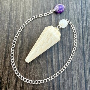 Peut inclure: Un pendule avec une pointe en cristal facettée de couleur crème, reliée par une chaîne argentée à une perle d'améthyste violette et une perle blanche. Le pendule est conçu pour la divination et les pratiques spirituelles.