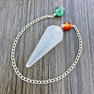 May include: A clear crystal pendulum with a silver chain and two colorful beads. The pendulum is cone-shaped and hangs from a silver chain. One end of the chain has a green and blue speckled bead, and the other end has two orange and red beads.