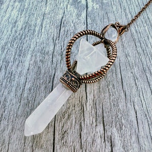 May include: A white crystal pendulum with a copper chain and a star-shaped crystal top. The pendulum is hanging from a wooden surface.